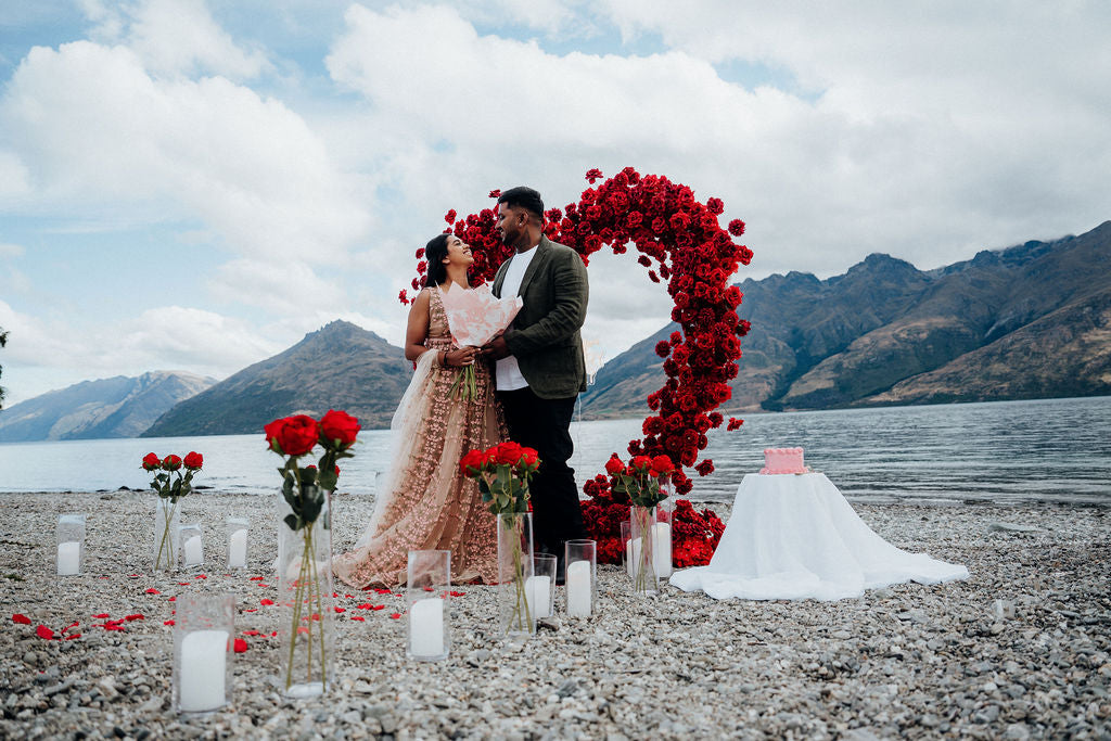 Couple standing under a heart-shaped arch of red flowers on a lakeside with mountains in the background