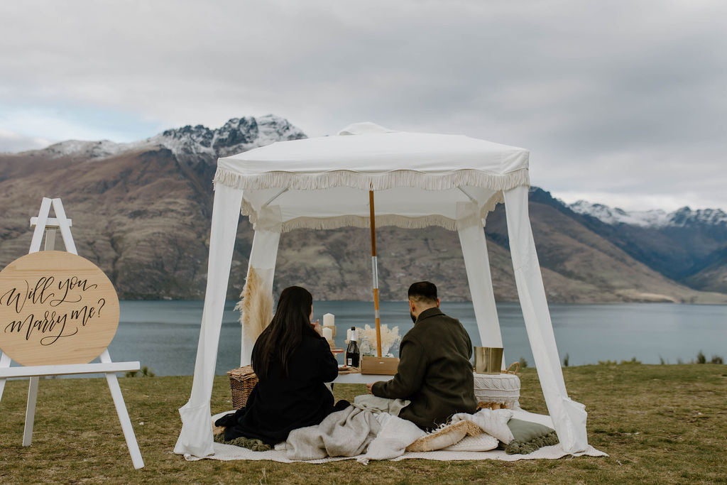 Couple sitting on cushions enjoying a picnic under a white canopy with a ‘Will you marry me?’ sign beside them.