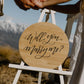 Two people holding hands with a wooden sign asking 'Will you marry me?' against a mountainous background.