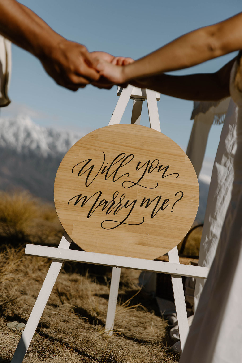 Two people holding hands with a wooden sign asking 'Will you marry me?' against a mountainous background.