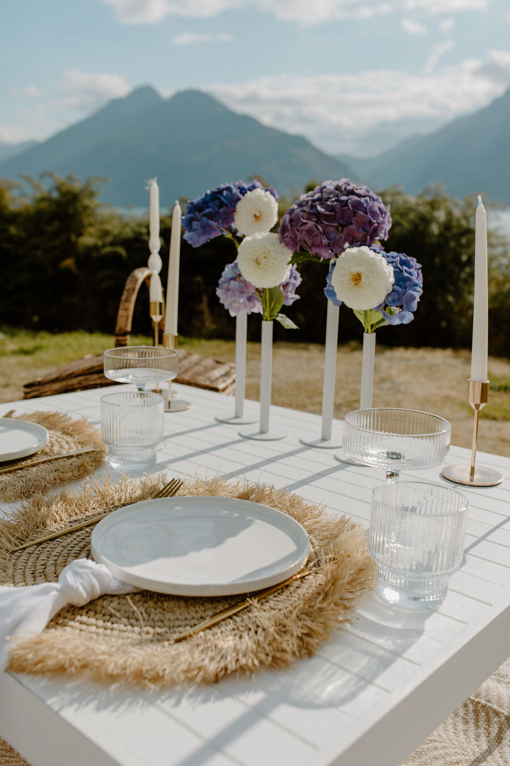 Elegant outdoor dining setup with flowers, plates, and glasses against a mountainous background.