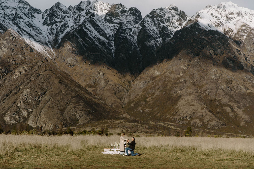 Man on one knee proposing to a woman in front of a romantic picnic in a field with mountains in the background