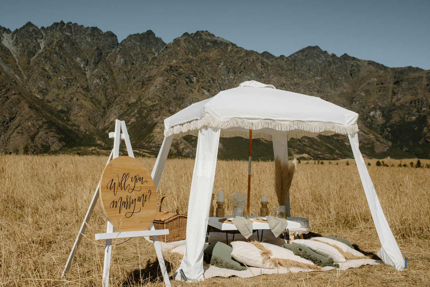 Romantic picnic setup with a table, pillows, white canopy and a ‘Will you marry me?’ sign in a mountain field.