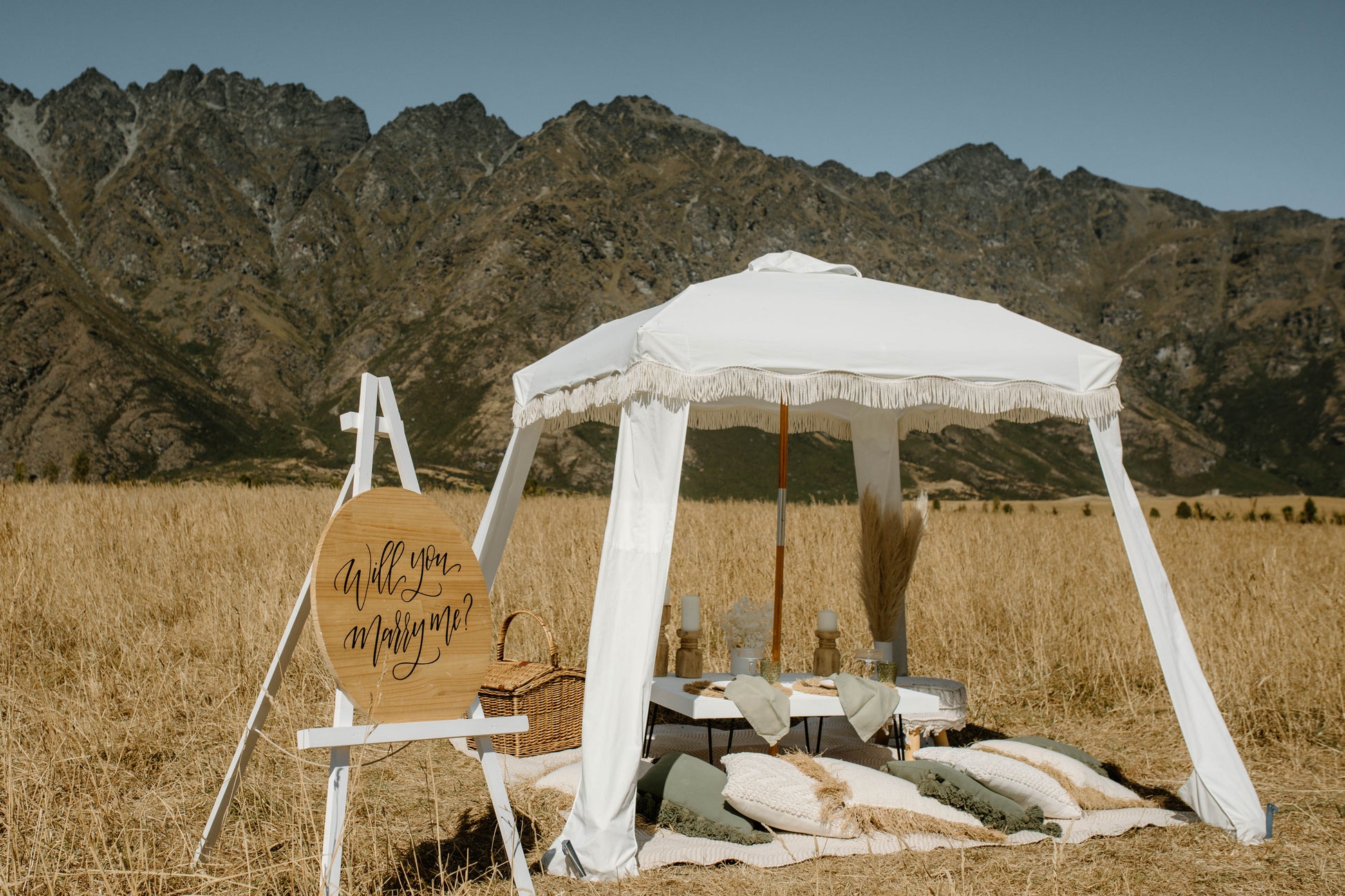 Romantic picnic setup with a table, pillows, white canopy and a ‘Will you marry me?’ sign in a mountain field.