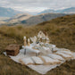 Romantic luxury picnic setup with table, chairs, and picnic basket on a blanket overlooking the mountains.