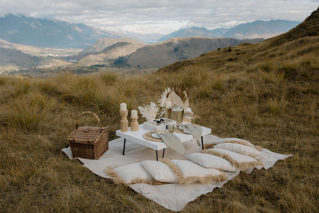 Romantic luxury picnic setup with table, chairs, and picnic basket on a blanket overlooking the mountains.