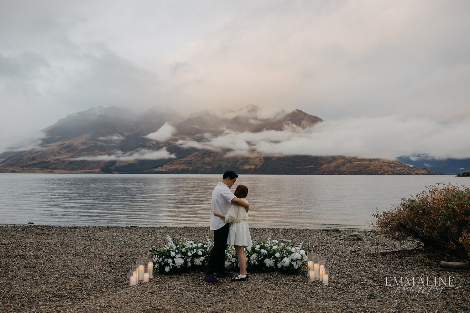 Couple embracing on a lakeside with candles and flowers, mountains in the background.