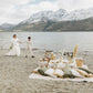Just-eloped couple holding hands by a romantic picnic on a beach with mountains in the background 