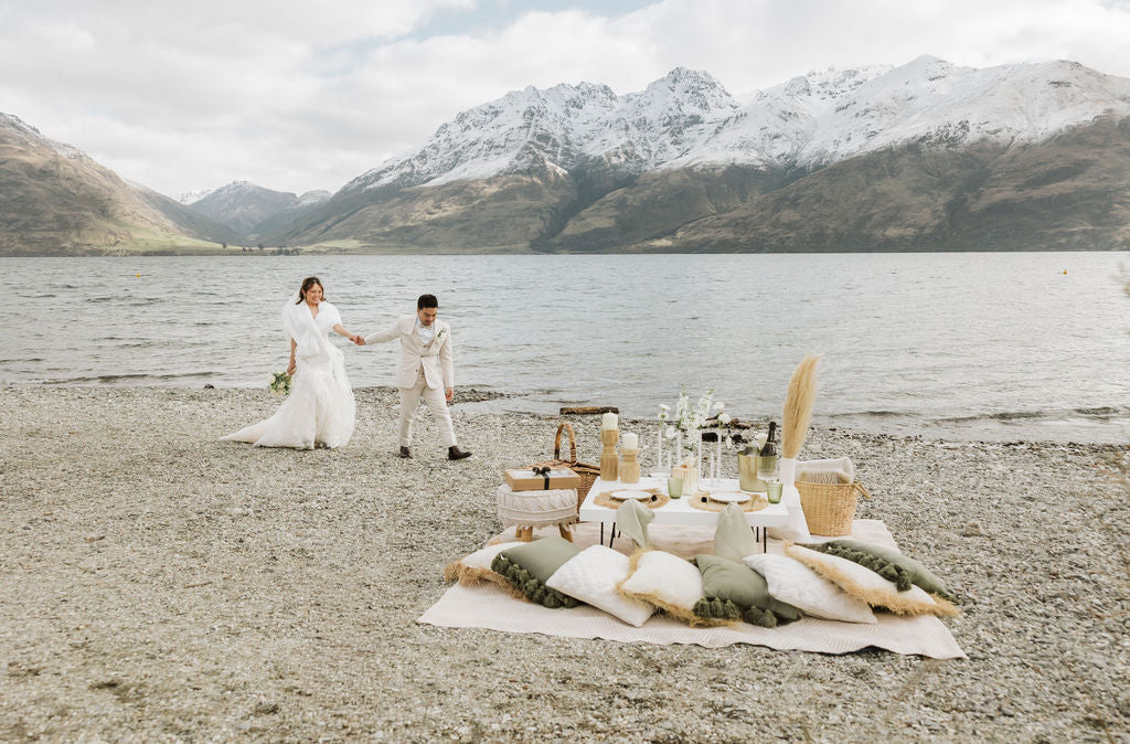 Just-eloped couple holding hands by a romantic picnic on a beach with mountains in the background 