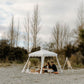 Two people enjoying a romantic picnic under a white canopy in a natural setting surrounded by trees and open space.