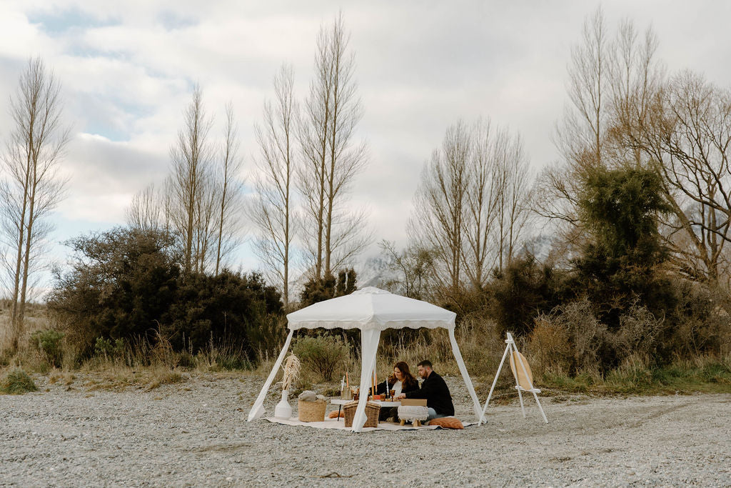 Two people enjoying a romantic picnic under a white canopy in a natural setting surrounded by trees and open space.