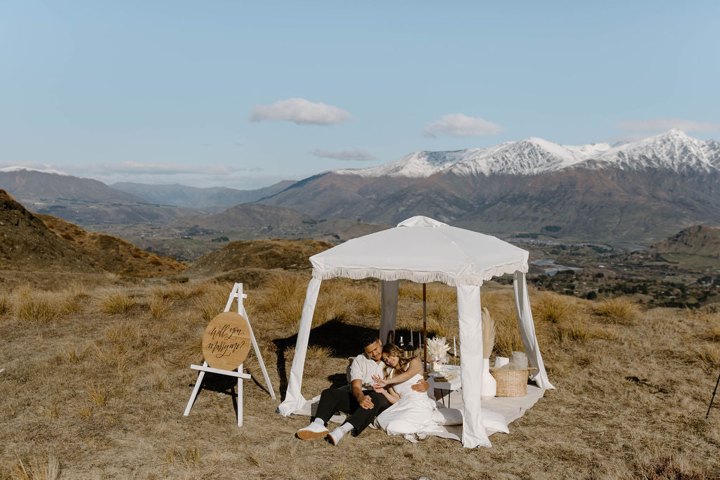 Newly engaged couple sitting under a white canopy with mountains in the background, admiring the engagement ring.